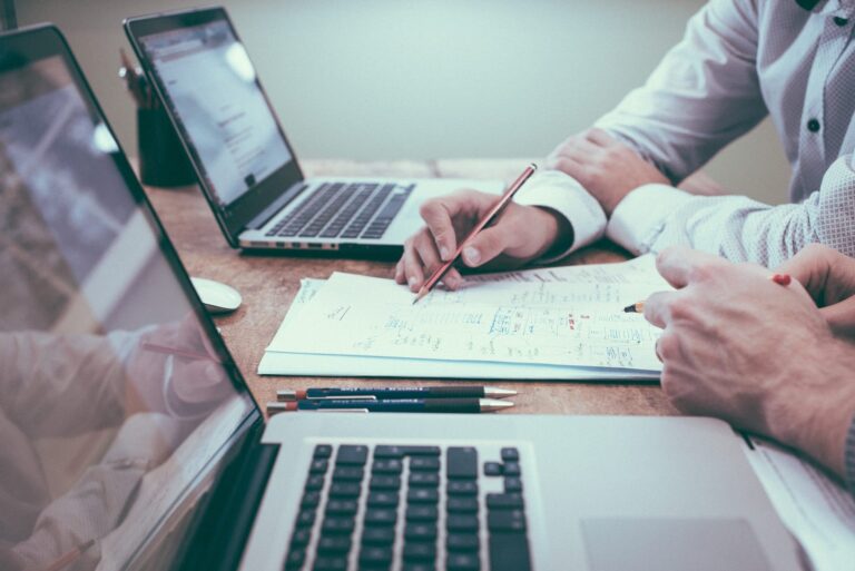 Two open computer on a table with with a contract in the middle. Two people start with pens in their hands looking at the contract.